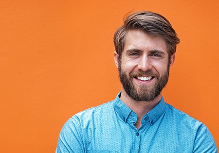 Een man met een baard glimlacht voor een oranje muur.